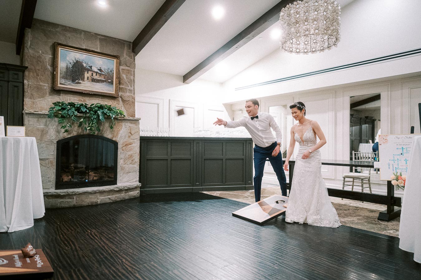 A bride and groom enjoying a game of cornhole indoors at their wedding reception, with a stone fireplace and elegant wood flooring in the background.