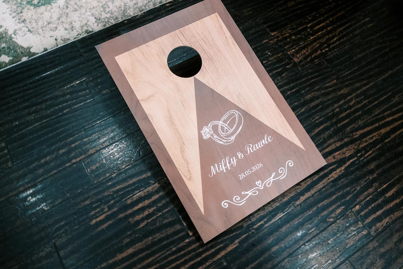 Wooden cornhole board with engraved wedding rings and names, set on a dark wooden floor.