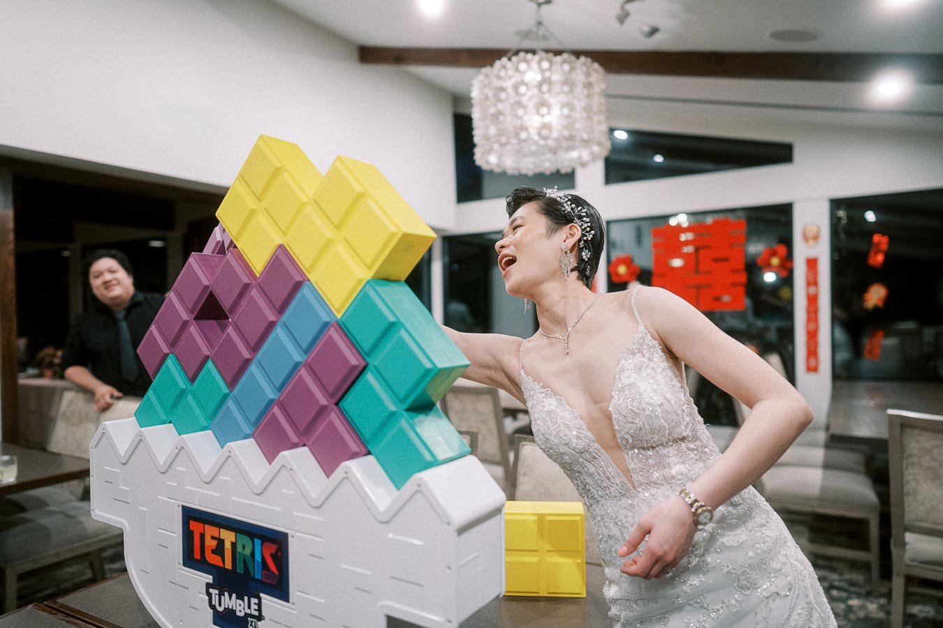 Bride playing with large Tetris pieces at a wedding reception, wearing a white lace gown and headpiece, with vibrant colors in the game and festive decorations in the background.