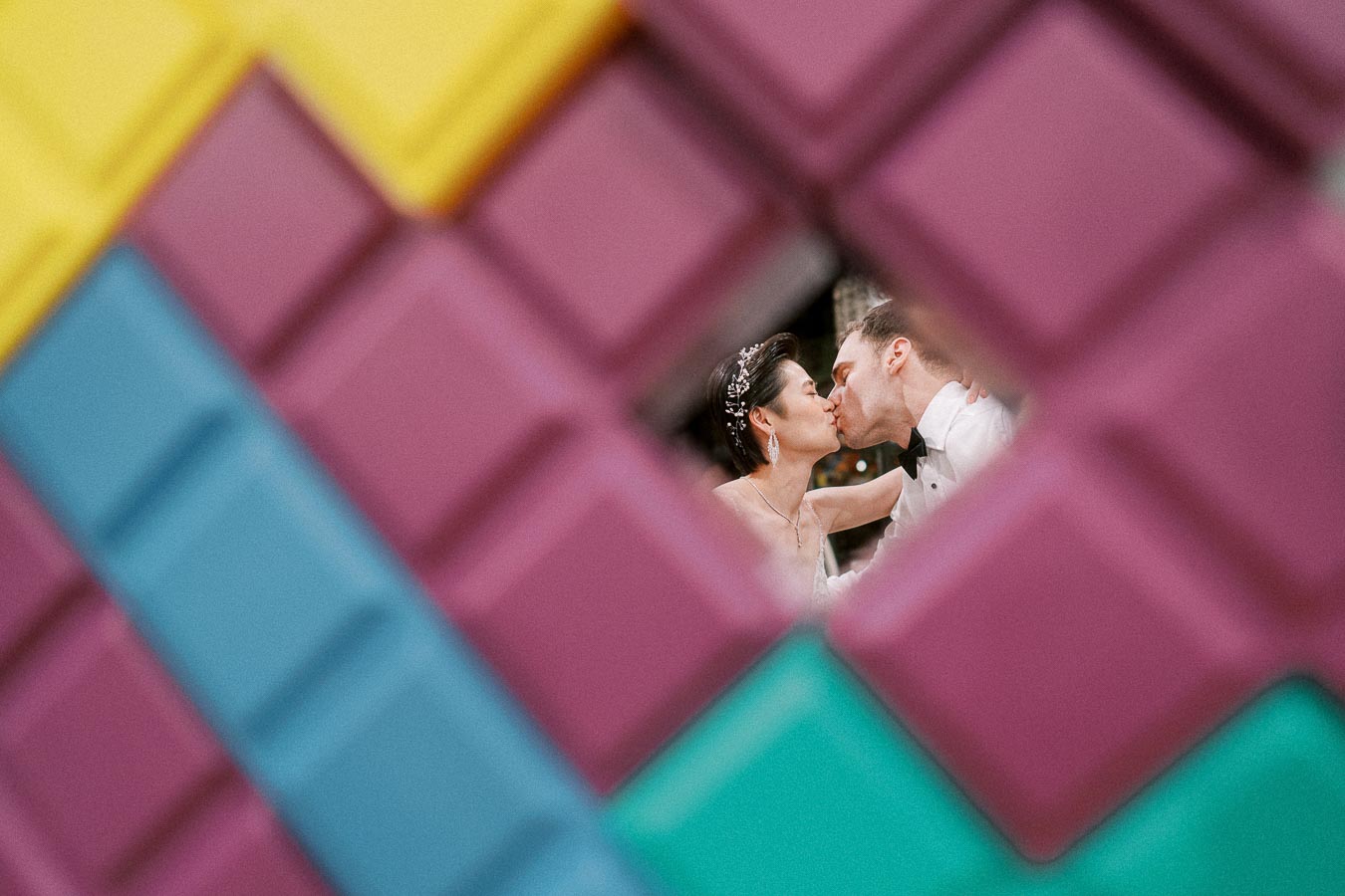 A creative and colorful wedding photograph featuring a couple kissing, captured through a vibrant geometric frame with yellow, blue, and purple blocks.