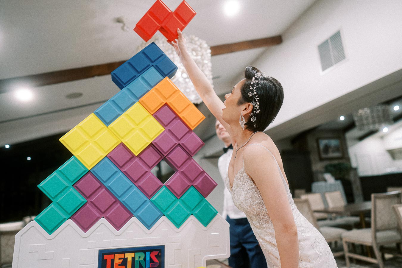 Woman in formal attire reaching to place a red Tetris block on top of a large colorful Tetris display at an indoor event.