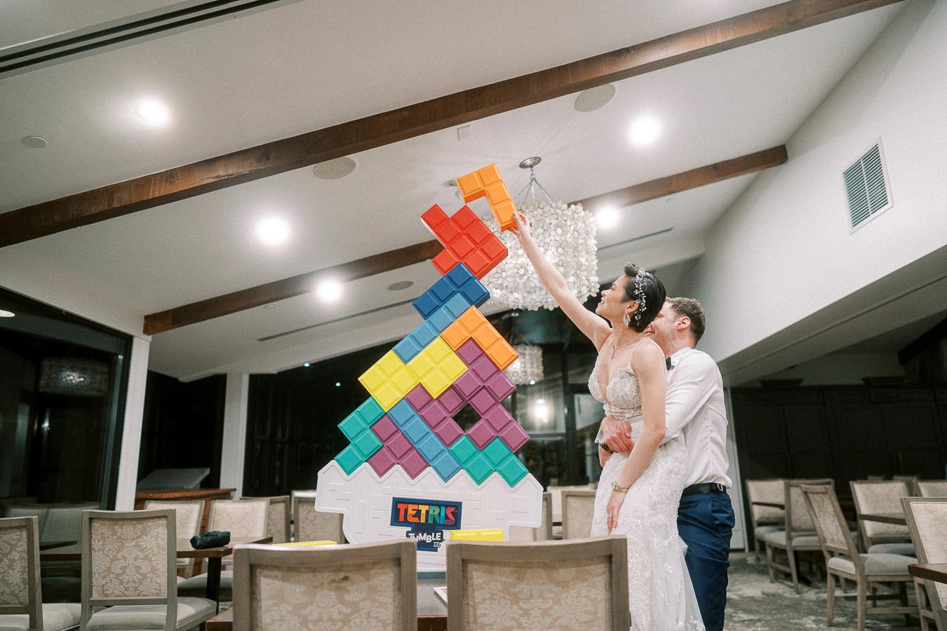 Bride and groom playing giant Tetris game at wedding reception, with the bride in a white gown placing a block while the groom supports her. Featuring colorful blocks in an elegant indoor setting.