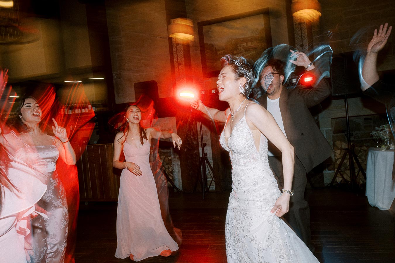 A group of people dancing energetically at a wedding reception, with the bride in a sparkling gown at the center, surrounded by colorful lights and a festive atmosphere.