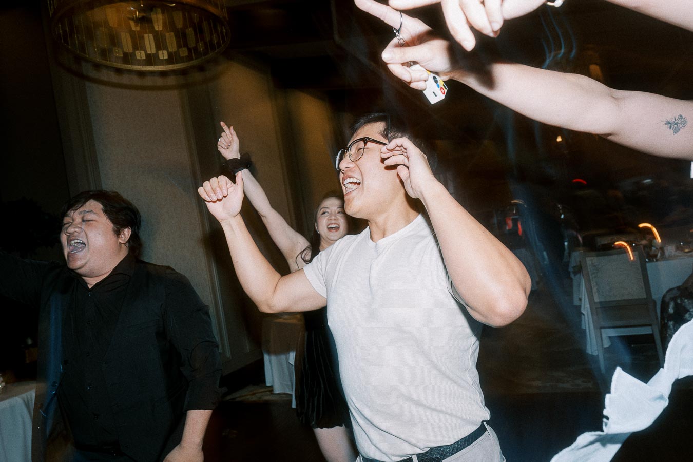 A group of people joyfully dancing at a lively indoor event, with one person in glasses and a white t-shirt raising their arms in celebration.