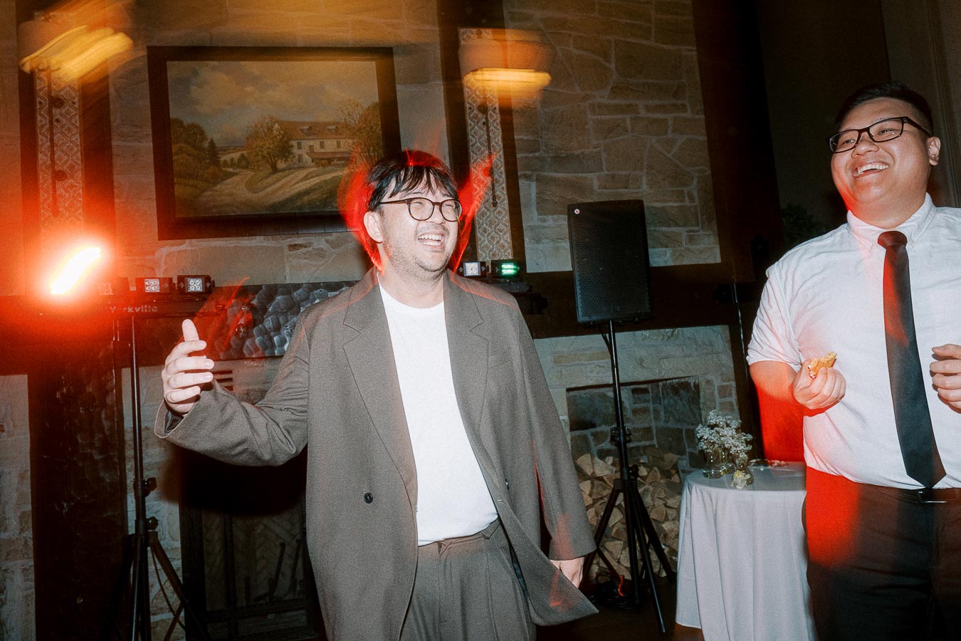 Two men laughing and enjoying a vibrant, warmly lit gathering in an indoor setting with stone walls and art décor. The atmosphere is lively and festive, featuring a blend of natural and artificial lighting, enhancing the joyful ambiance.