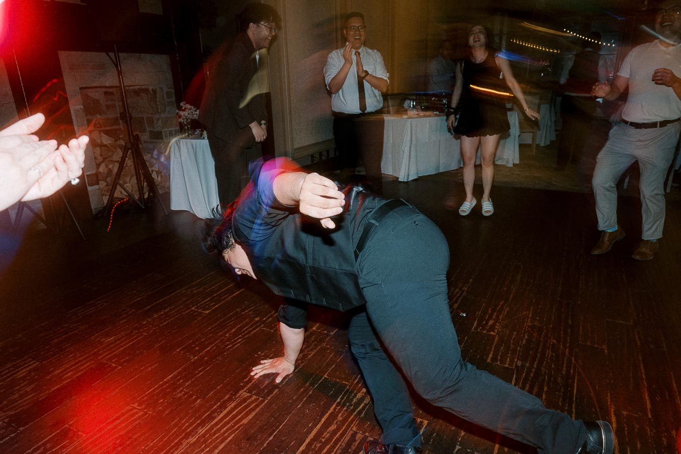 A person dancing energetically on a dimly lit floor at a lively party, surrounded by cheering friends.