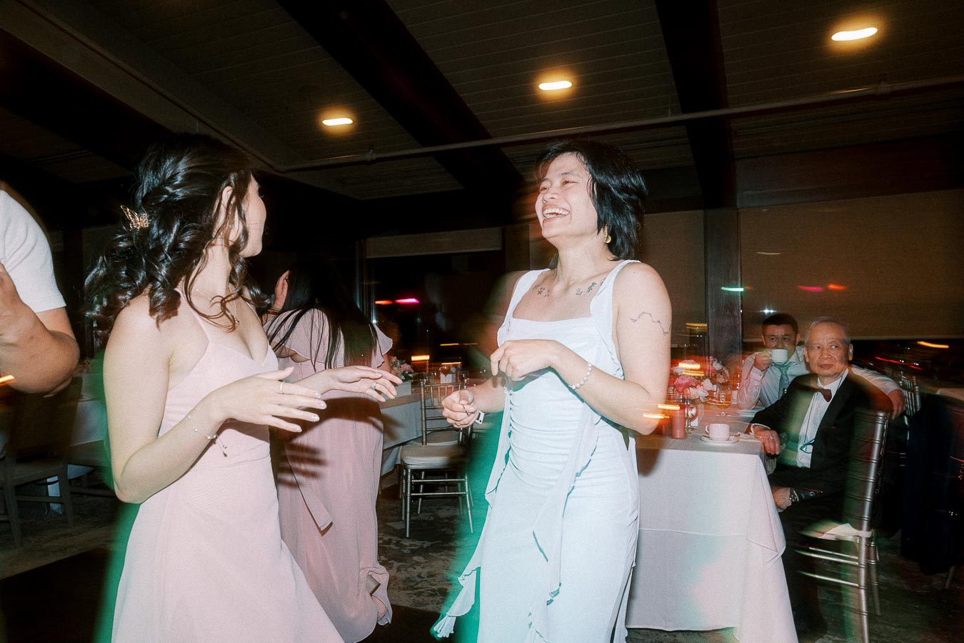 Two women in elegant dresses laughing and dancing at a lively indoor event, with seated guests enjoying the evening in the background.