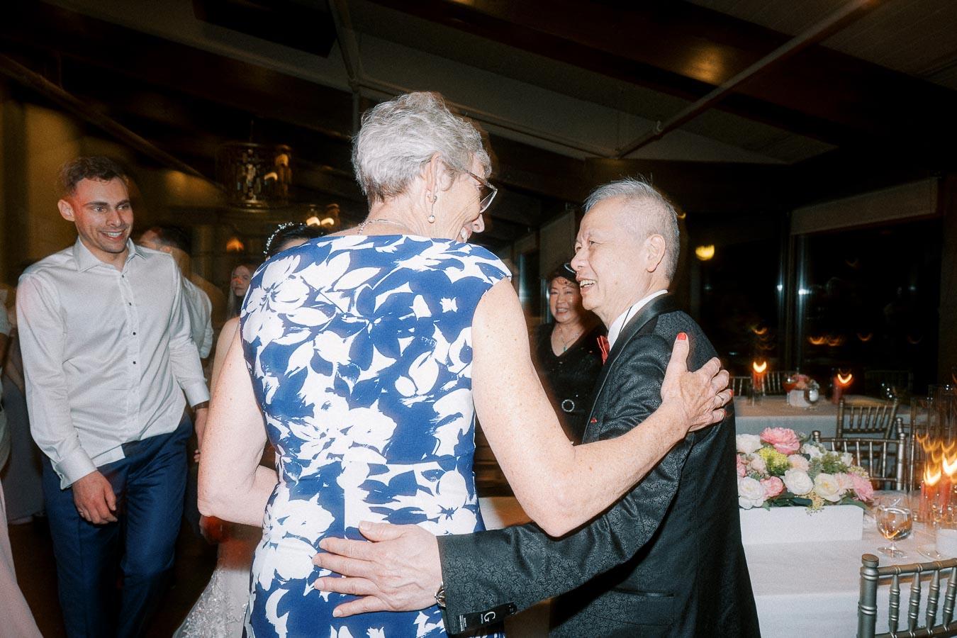Elderly guests smiling and embracing at a lively wedding reception, with elegantly decorated tables and other attendees enjoying the celebration in the background.