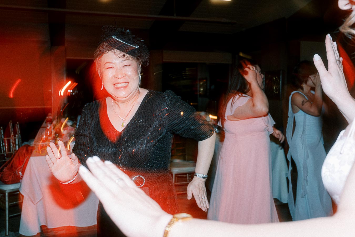 A joyful woman in a black dress dances at a lively party, surrounded by friends in colorful attire, with a vibrant red light adding to the festive atmosphere.