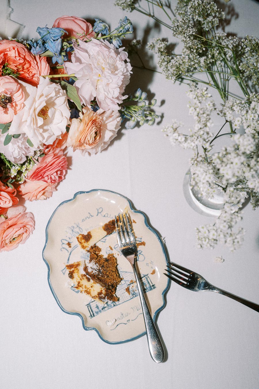 A delicate floral bouquet featuring pink, white, and blue flowers beside a vintage-patterned plate with remnants of a cake slice and two silver forks, elegantly set on a white tablecloth.