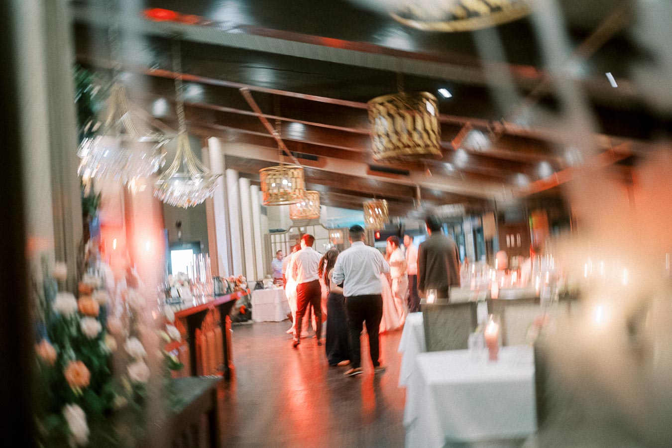 Blurred image of an indoor event with people socializing in a warmly lit, elegantly decorated venue, featuring chandeliers and floral arrangements.