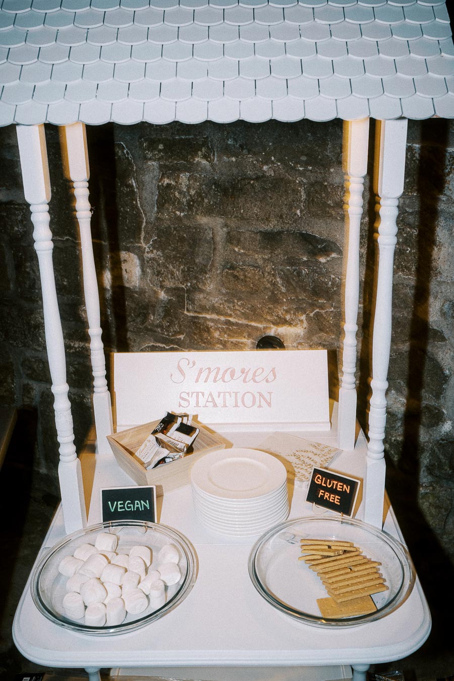 S'mores station setup with vegan marshmallows and gluten-free graham crackers on a white cart against a rustic stone wall.