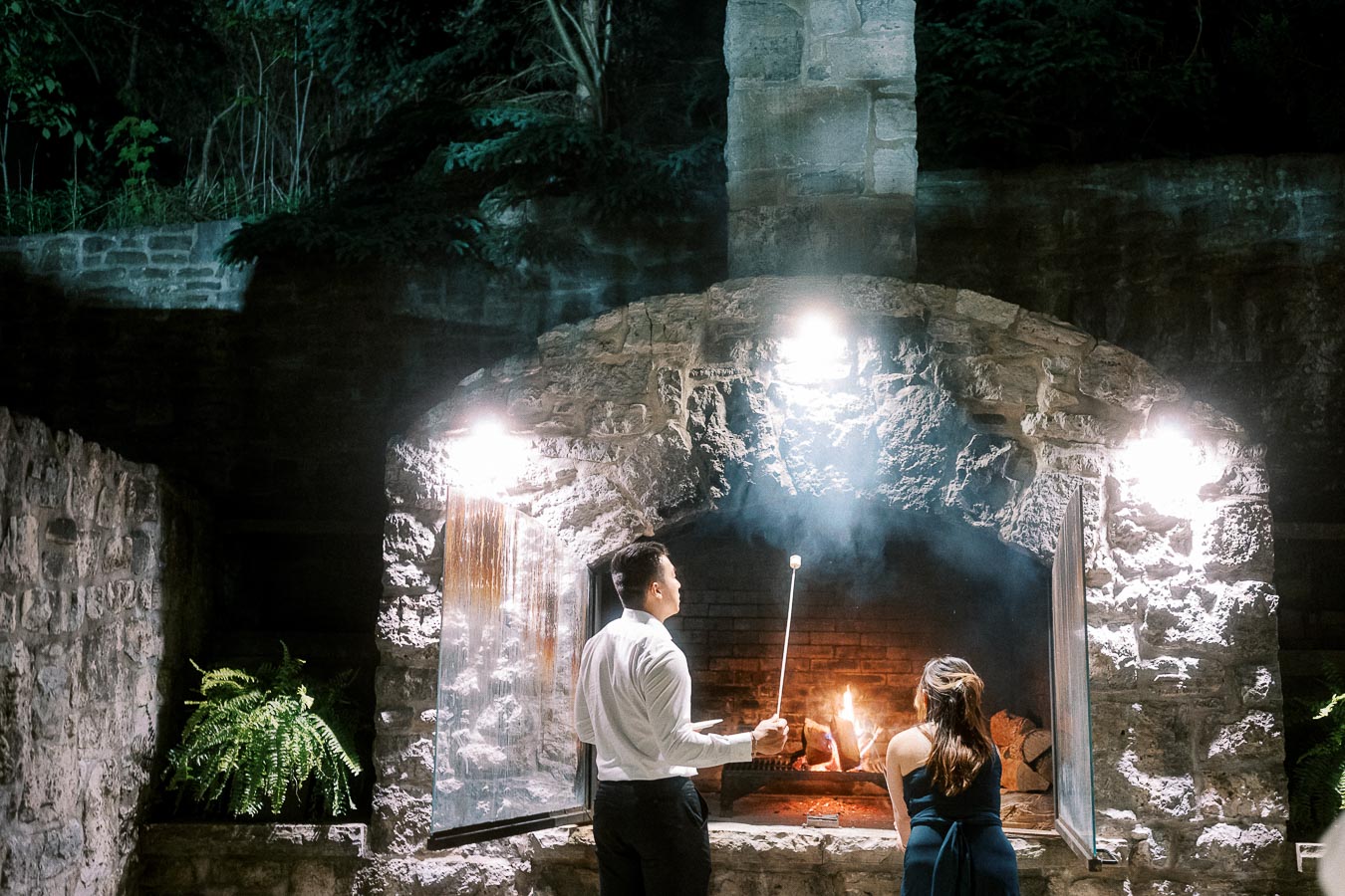 A couple roasting marshmallows over an outdoor stone fireplace at night, surrounded by lush greenery and illuminated by soft lighting.