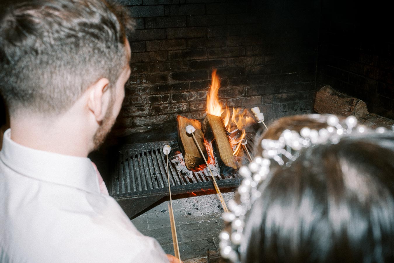 A couple roasting marshmallows over a cozy fireplace, with logs burning brightly in a rustic brick setting.