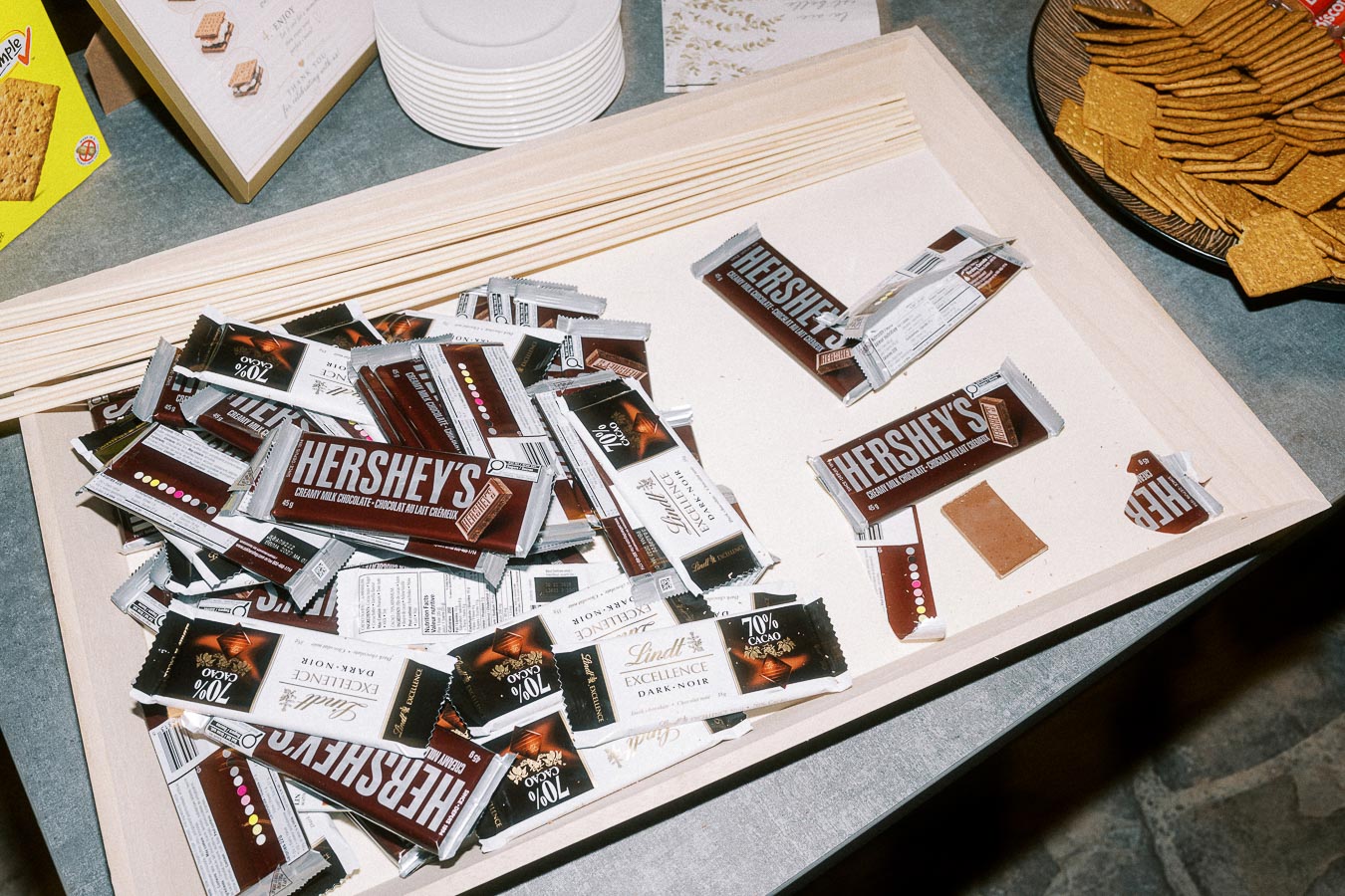 A wooden tray filled with assorted chocolate bars, including Hershey's and Lindt, set on a table next to graham crackers, ready for making s'mores.