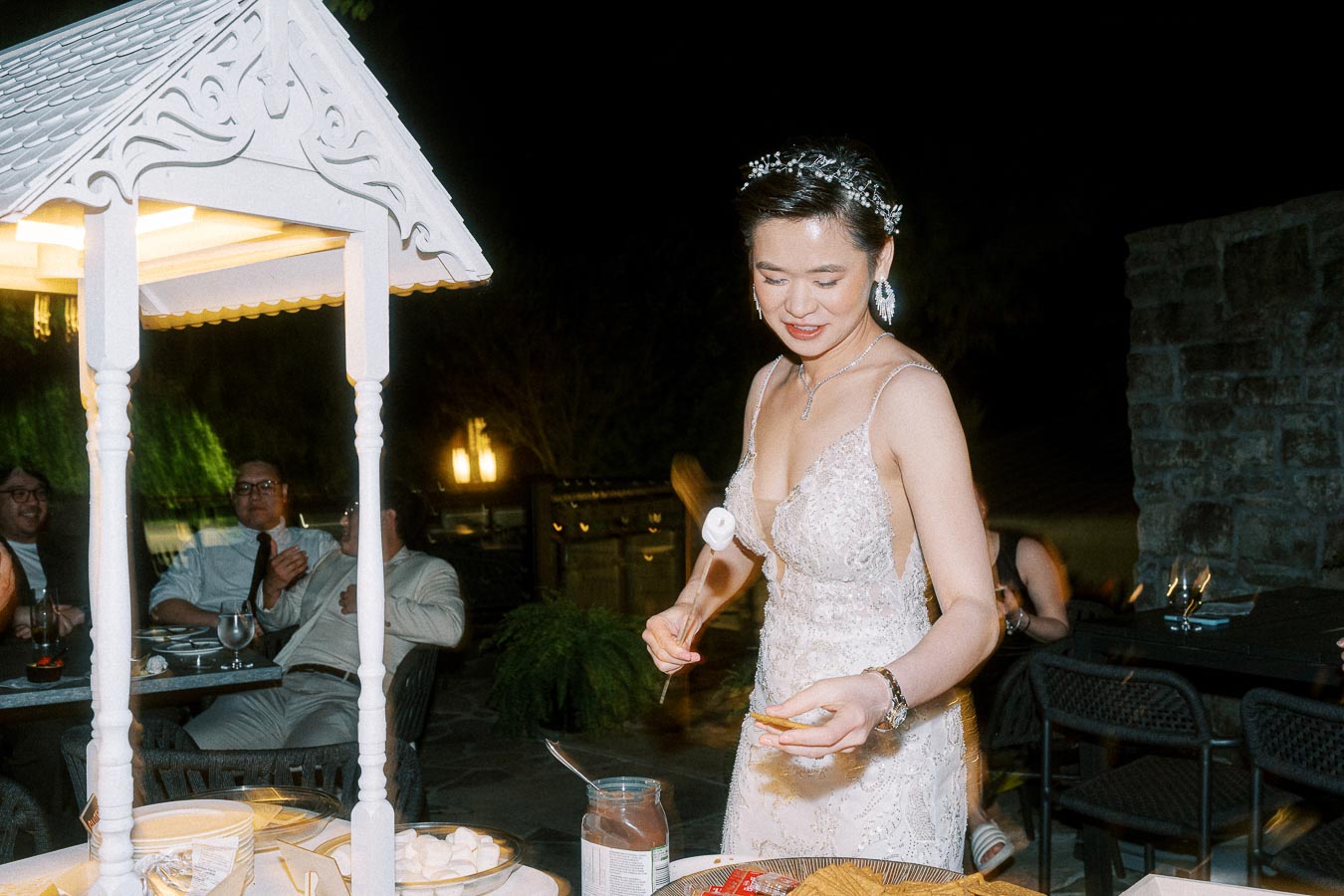 Elegant bride in a beautiful lace wedding dress prepares a sweet treat at a stylish outdoor wedding reception, surrounded by glowing lights and guests in evening attire.