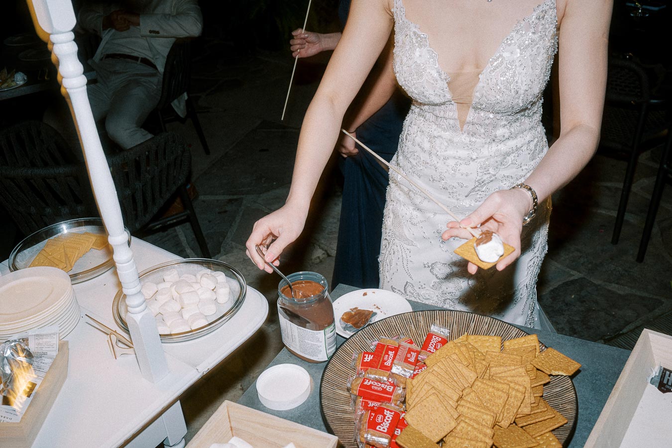 A person in an elegant white lace dress prepares a s'more with graham crackers, marshmallows, and chocolate at an outdoor party setup. A variety of s'mores ingredients are displayed on a table, creating a festive and inviting atmosphere.