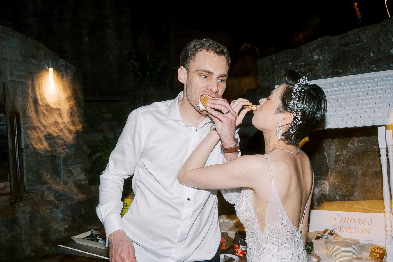 Bride and groom enjoying s'mores together at their wedding reception, with a rustic stone wall and softly lit ambiance in the background.
