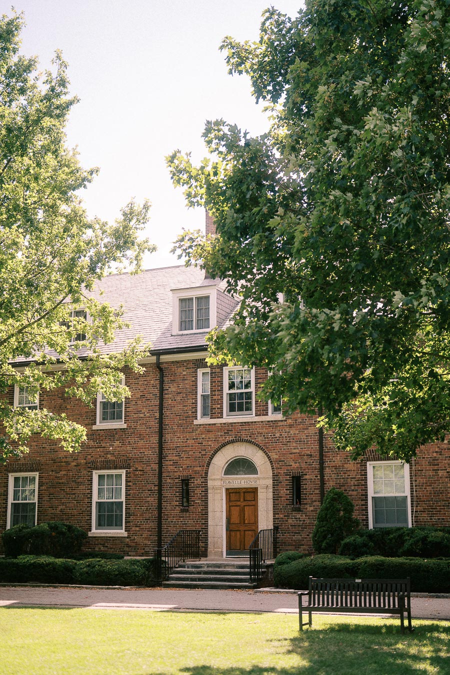 Brick campus building framed by leafy trees, with an arched doorway, front steps, lawn, and bench.