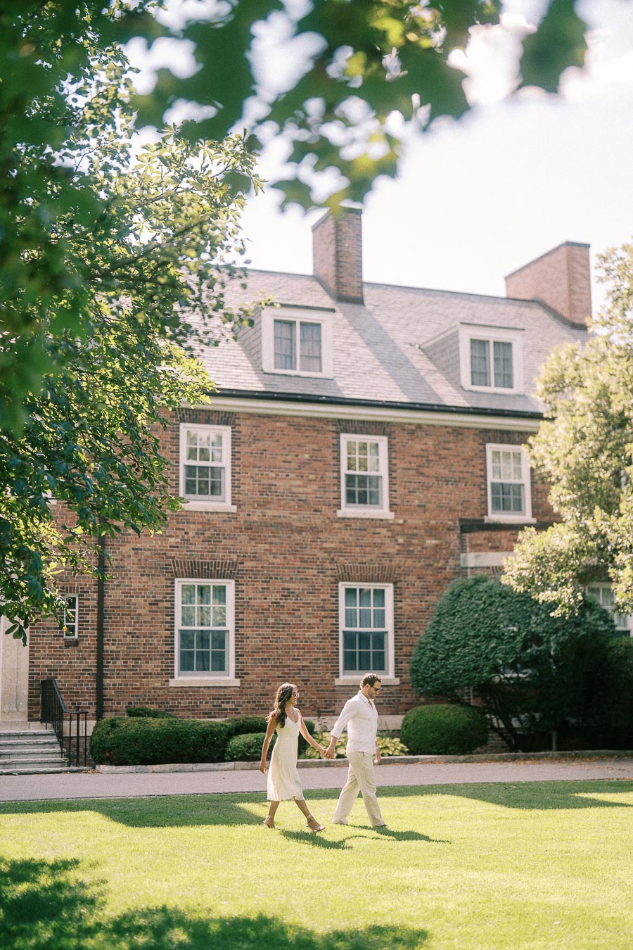 Rachel and Matt walk hand in hand across a sunny lawn in front of a brick school building in Aurora.