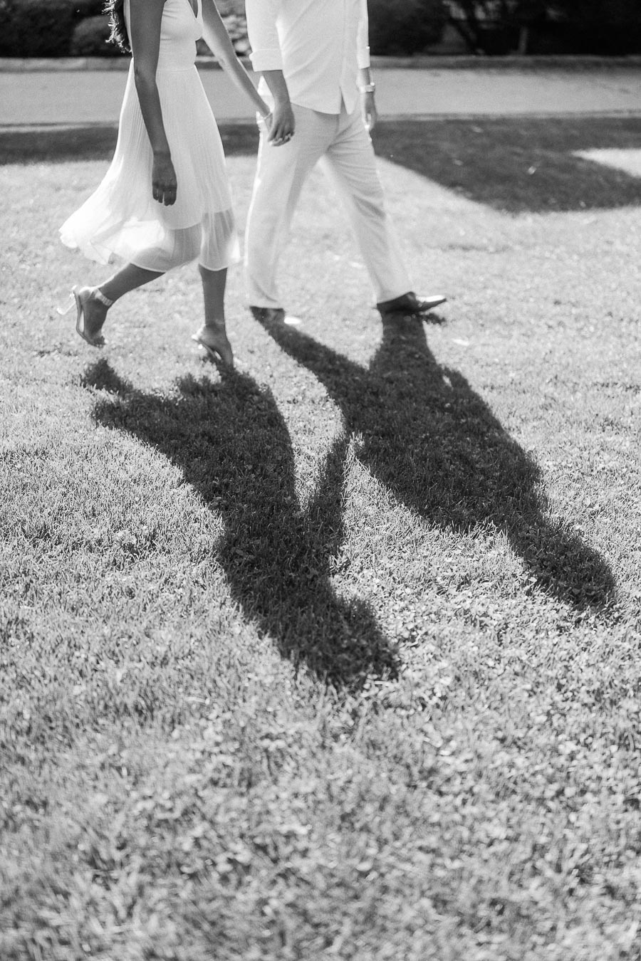 Rachel and Matt walk hand in hand across sunlit grass, their long shadows stretching toward the camera.
