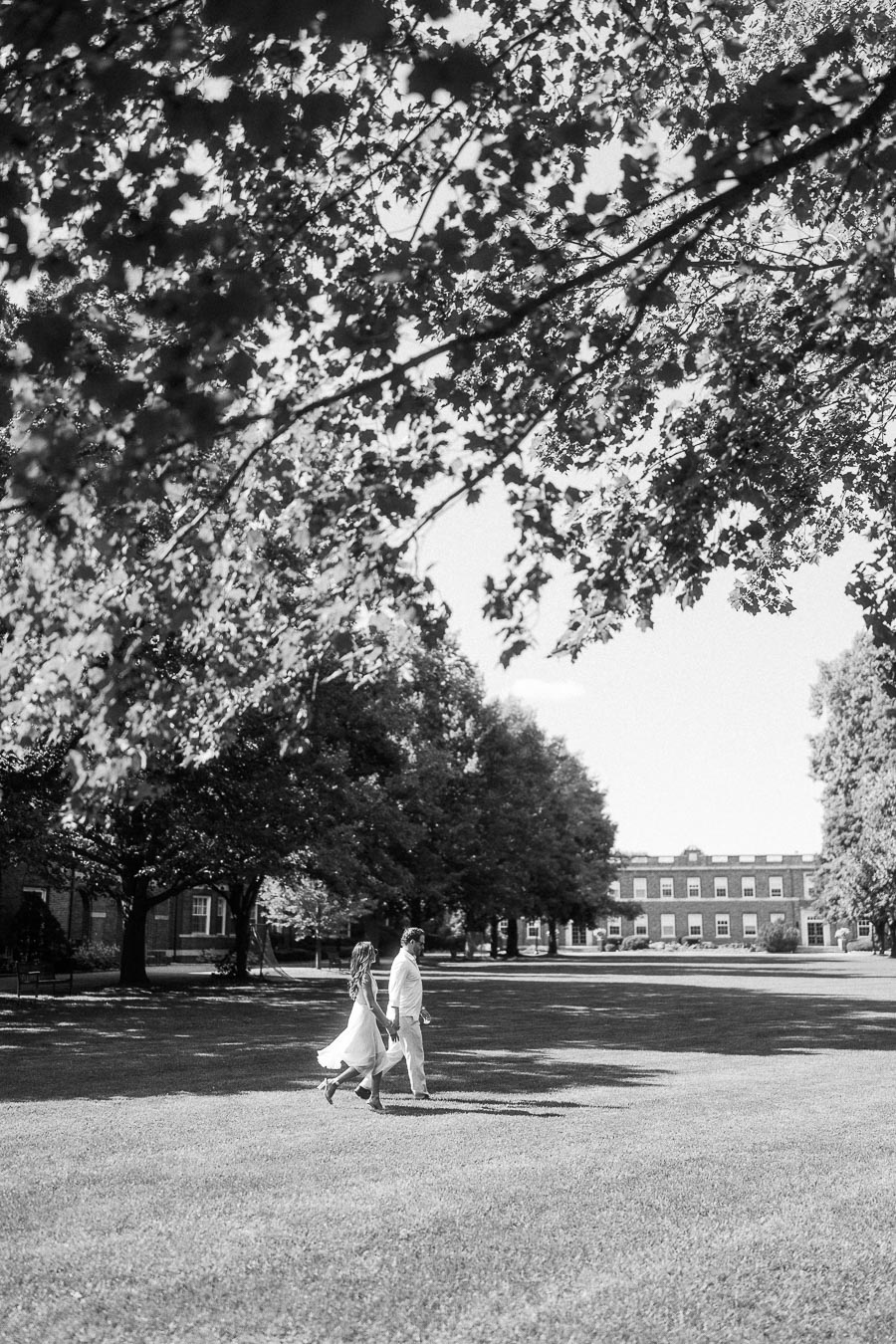 Rachel and Matt walk hand in hand across a broad campus lawn beneath leafy trees in black and white.