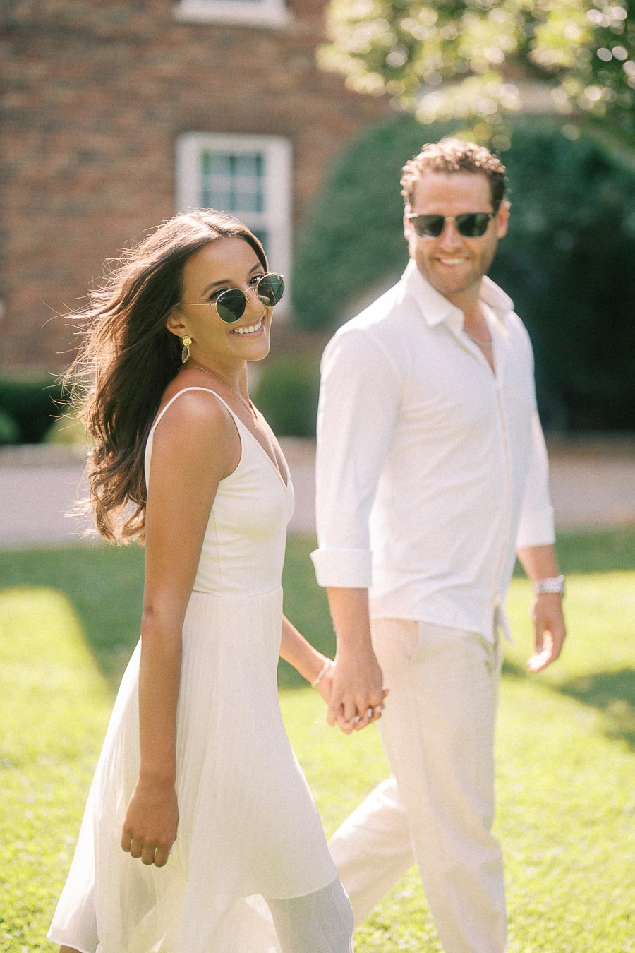 Rachel and Matt hold hands while walking on a sunny lawn, both smiling and wearing sunglasses.