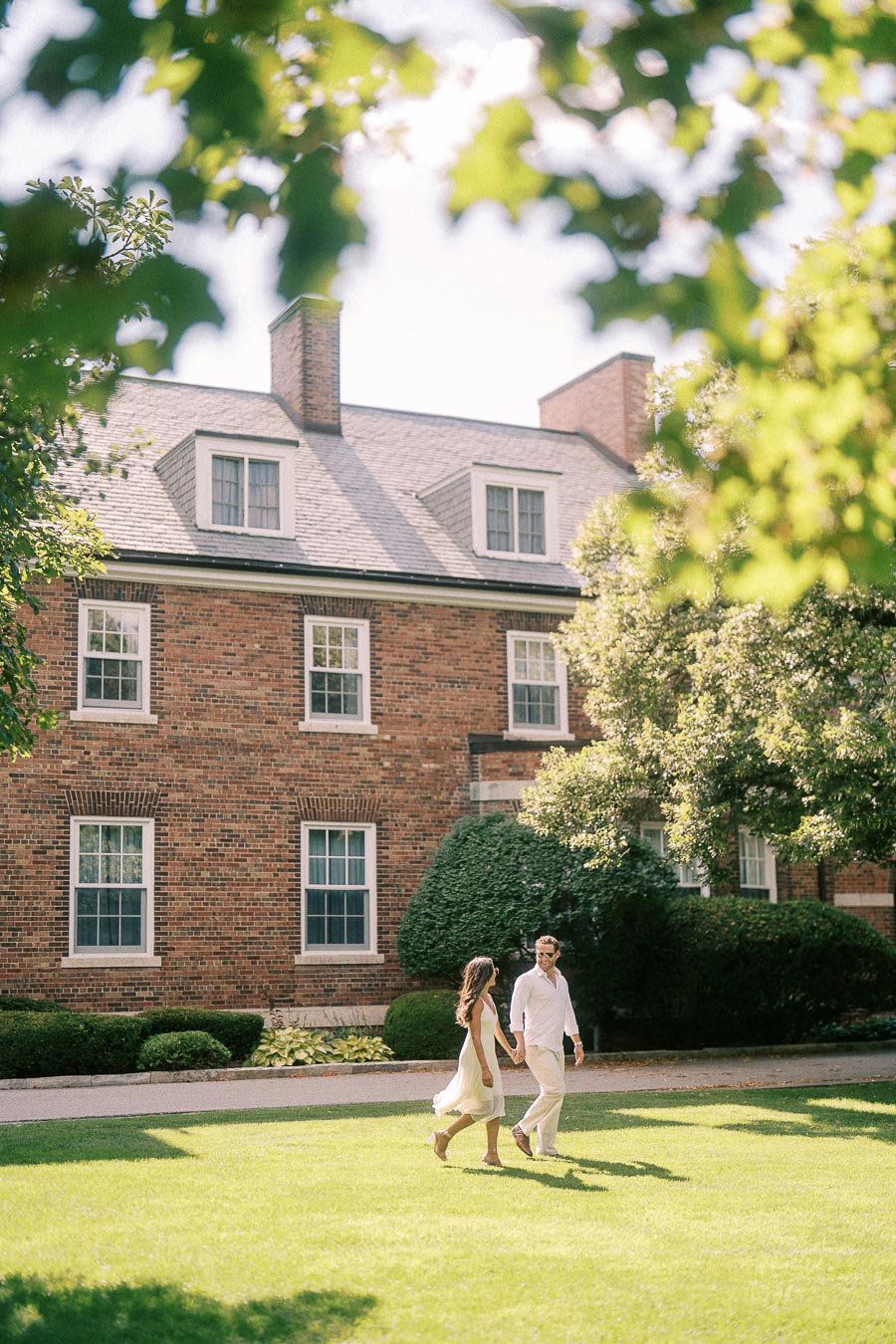 Rachel and Matt hold hands while walking across a sunny lawn beside a brick school building in Aurora.