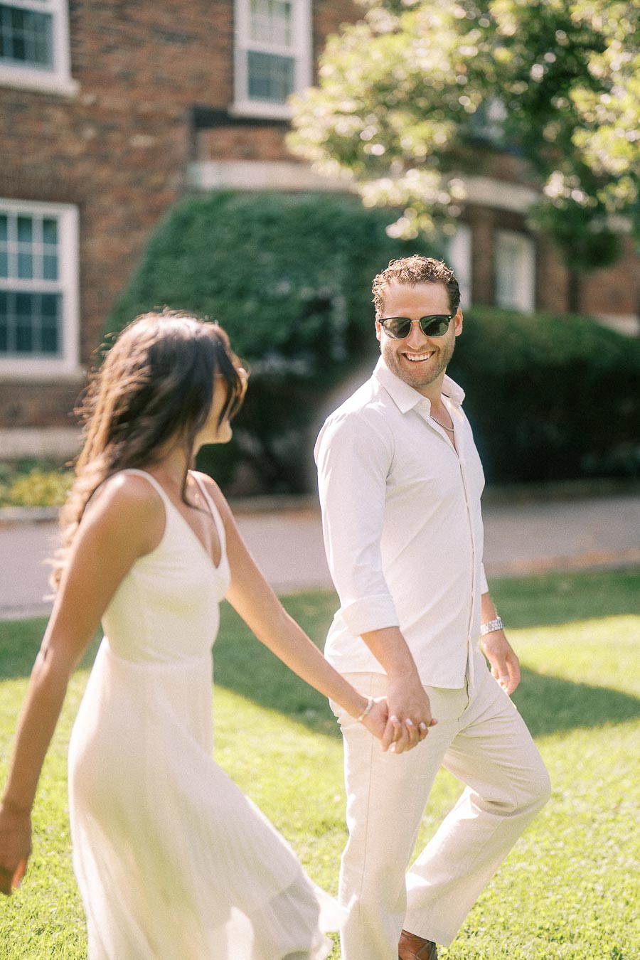 Rachel and Matt hold hands while walking across a sunny lawn in front of a brick building.