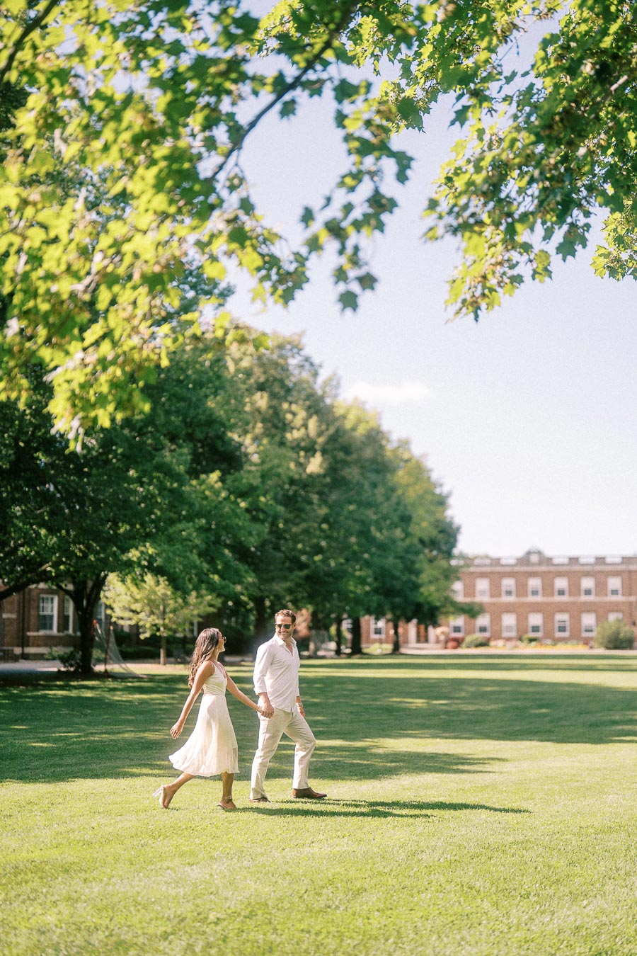 Rachel and Matt walk hand in hand across a sunny campus lawn under leafy trees in Aurora, Ontario.