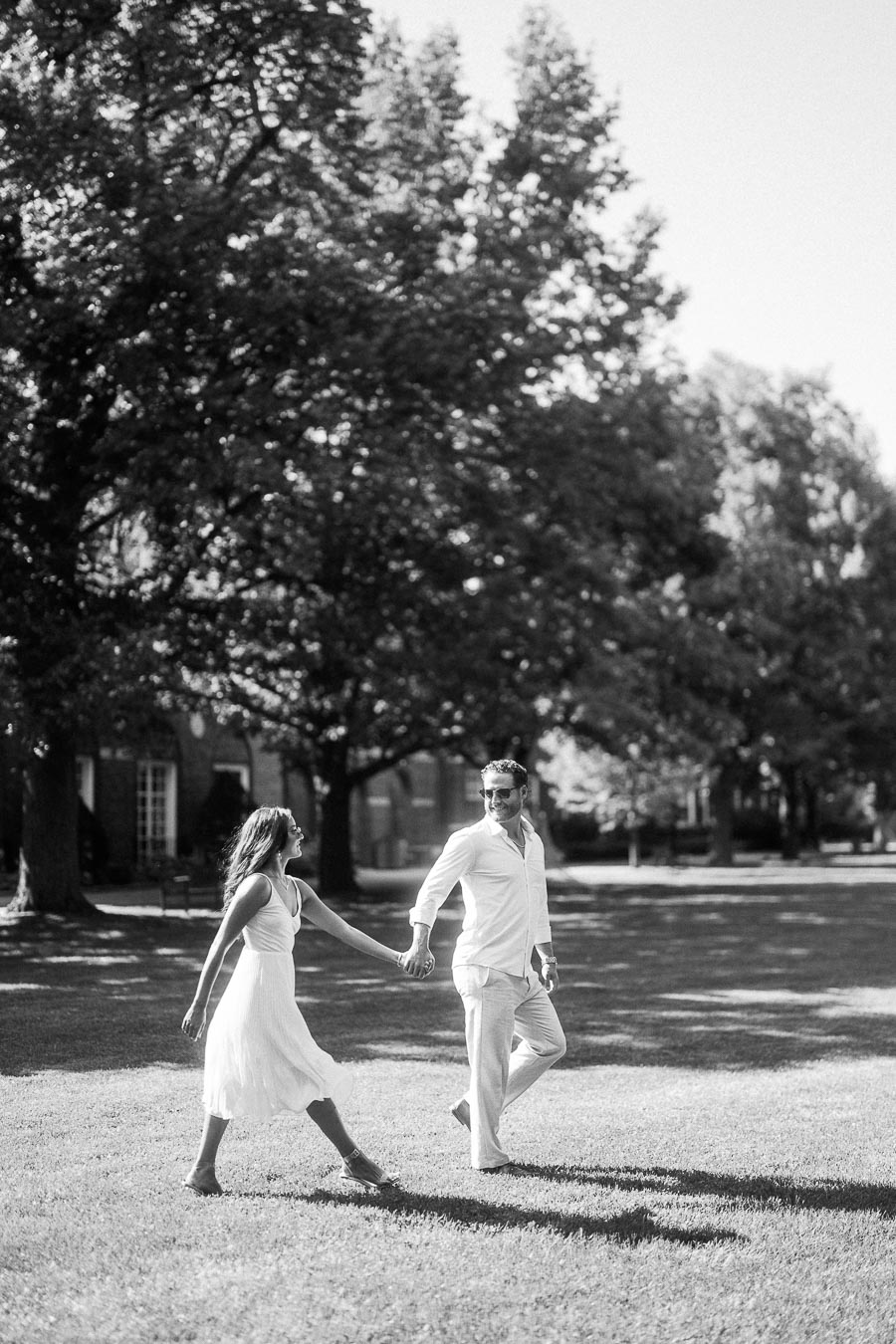 Rachel and Matt hold hands while walking across a sunny lawn beneath large trees in Aurora, Ontario.