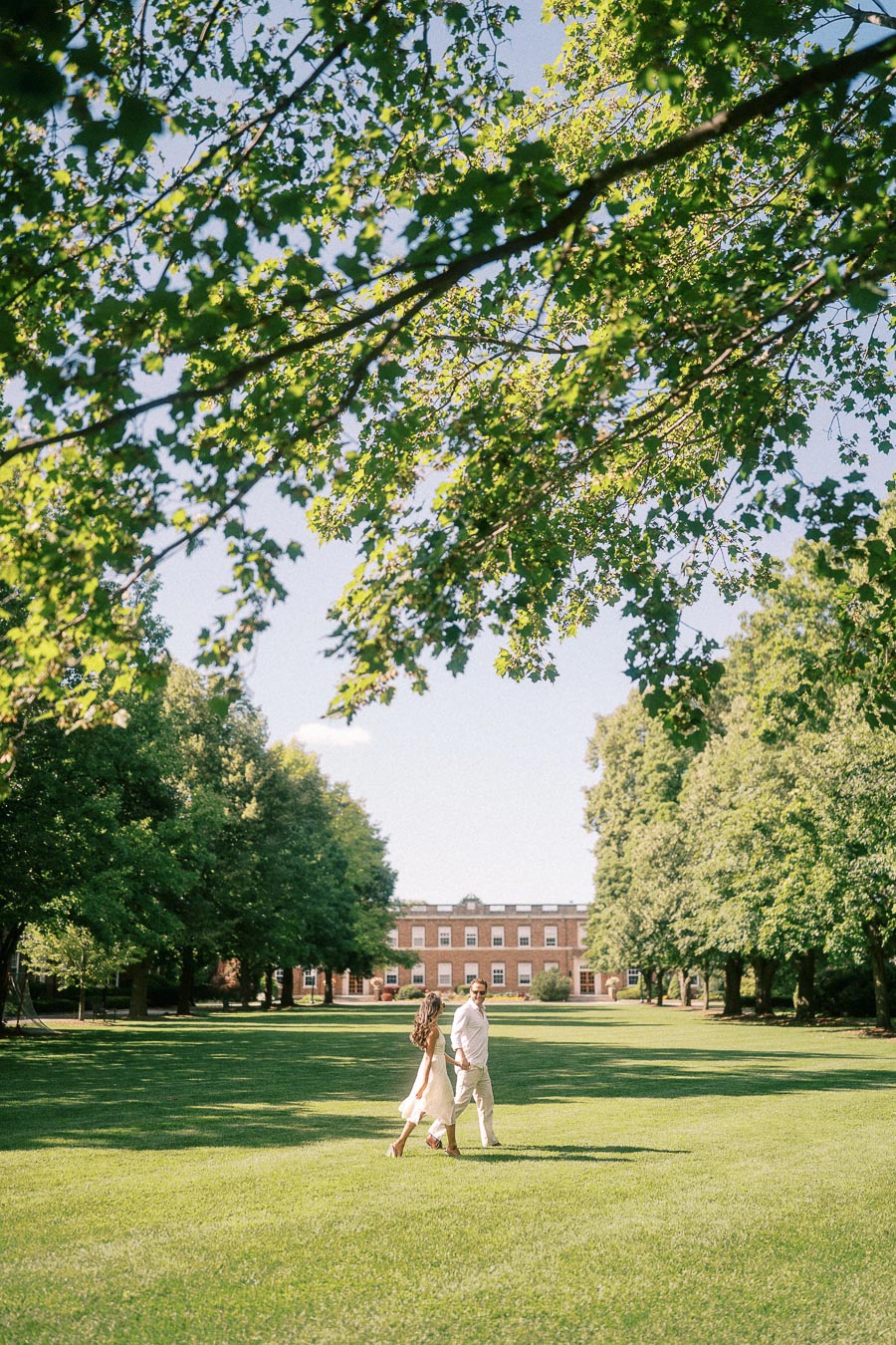Rachel and Matt walk hand in hand across a wide green campus lawn beneath leafy summer trees.