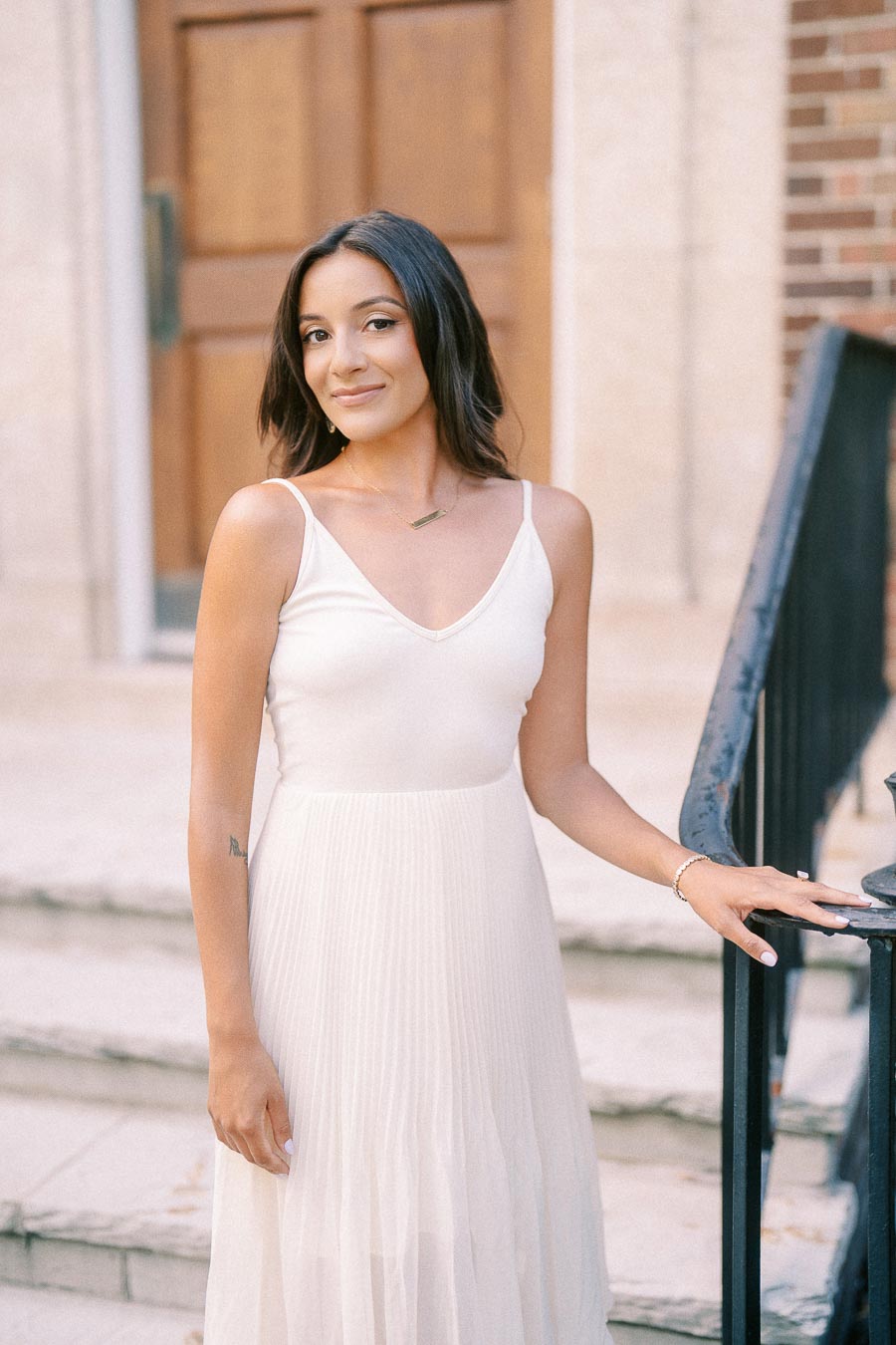 Rachel stands in a white dress beside a black railing and stone steps outside a brick building.