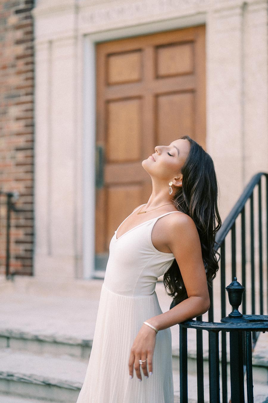 Rachel stands in a white dress beside a black railing, leaning back near a wooden doorway in Aurora.