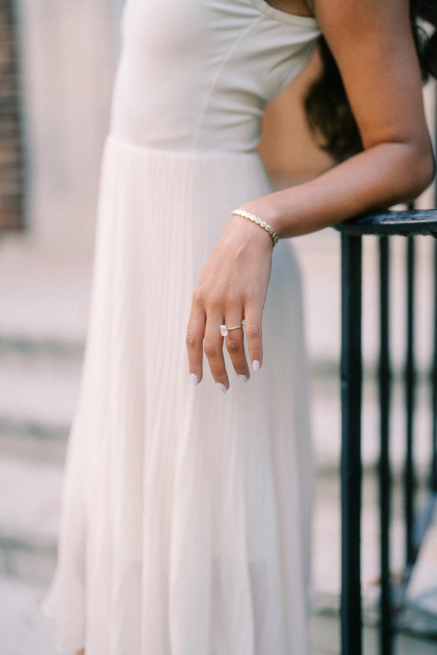 Rachel’s hand rests beside a black railing, showing her engagement ring and bracelet against a white dress.