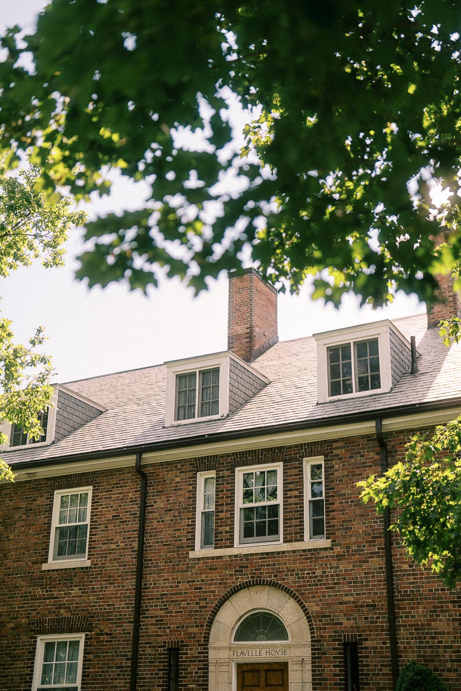 Brick Flavelle House facade with dormer windows and leafy tree branches overhead in bright summer light.