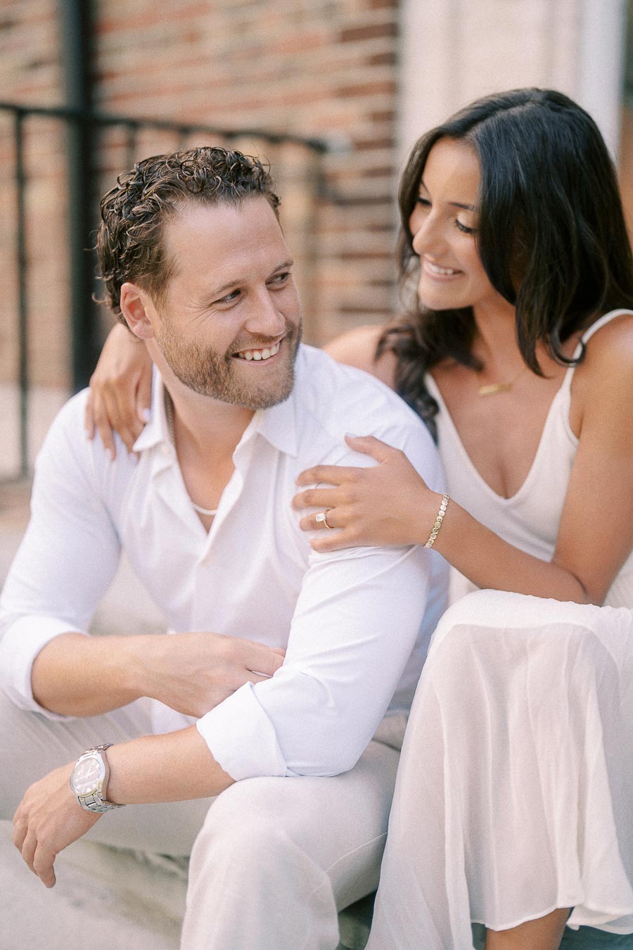 Rachel rests her hand on Matt’s shoulder as they sit together smiling in front of a brick wall.