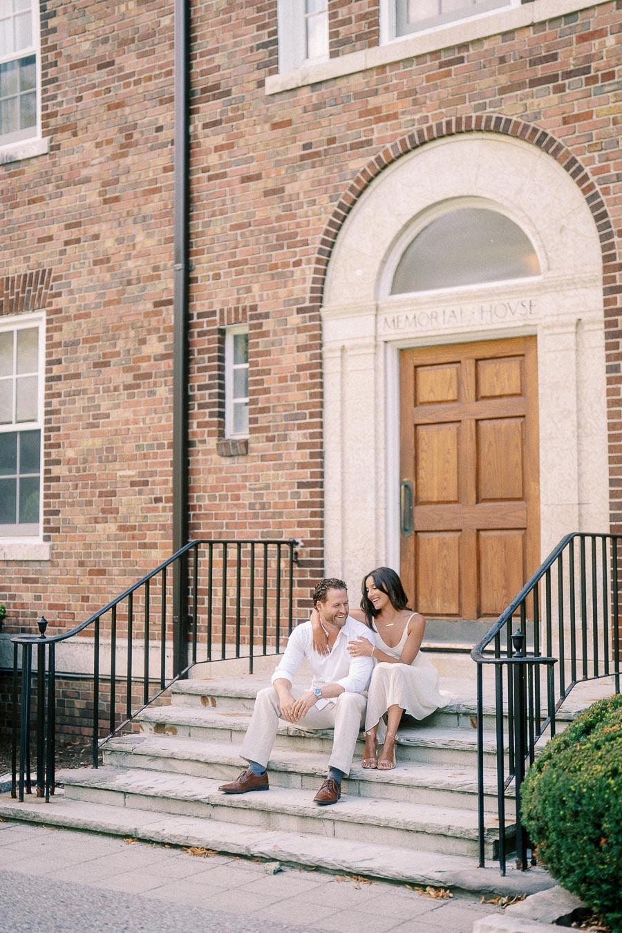 Rachel and Matt sit together on stone steps outside Memorial House, smiling beside a brick entrance.