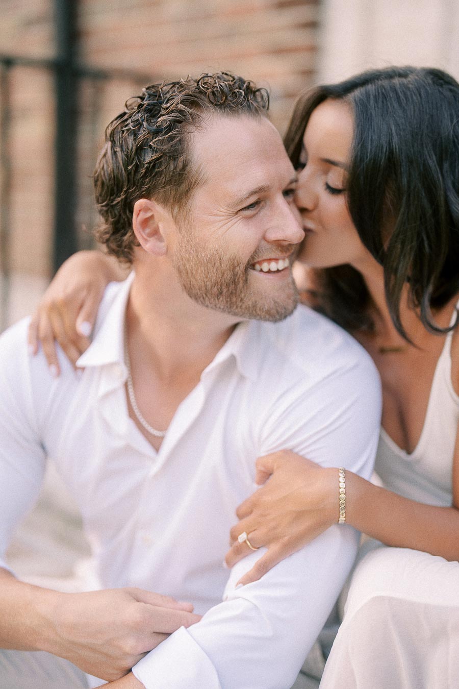 Rachel kisses Matt on the cheek as he smiles, seated close together in light outfits outdoors.