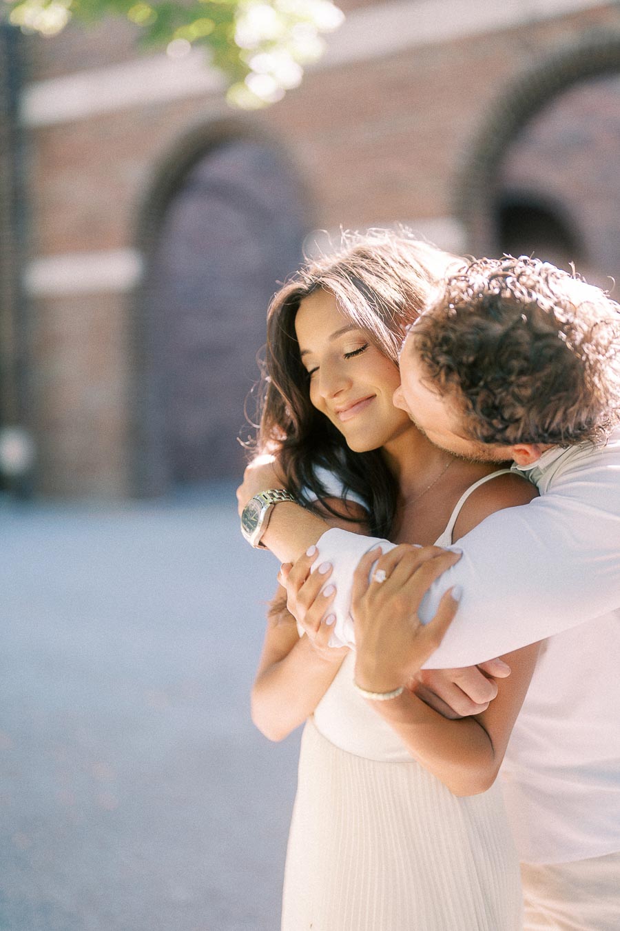 Matt hugs Rachel from behind and kisses her cheek in front of a sunlit brick building with arched windows.