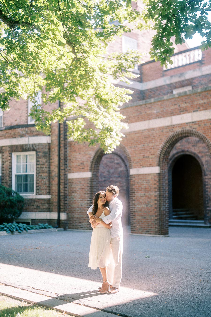 Rachel and Matt embrace in a sunlit courtyard beside a brick building with arched doorways.