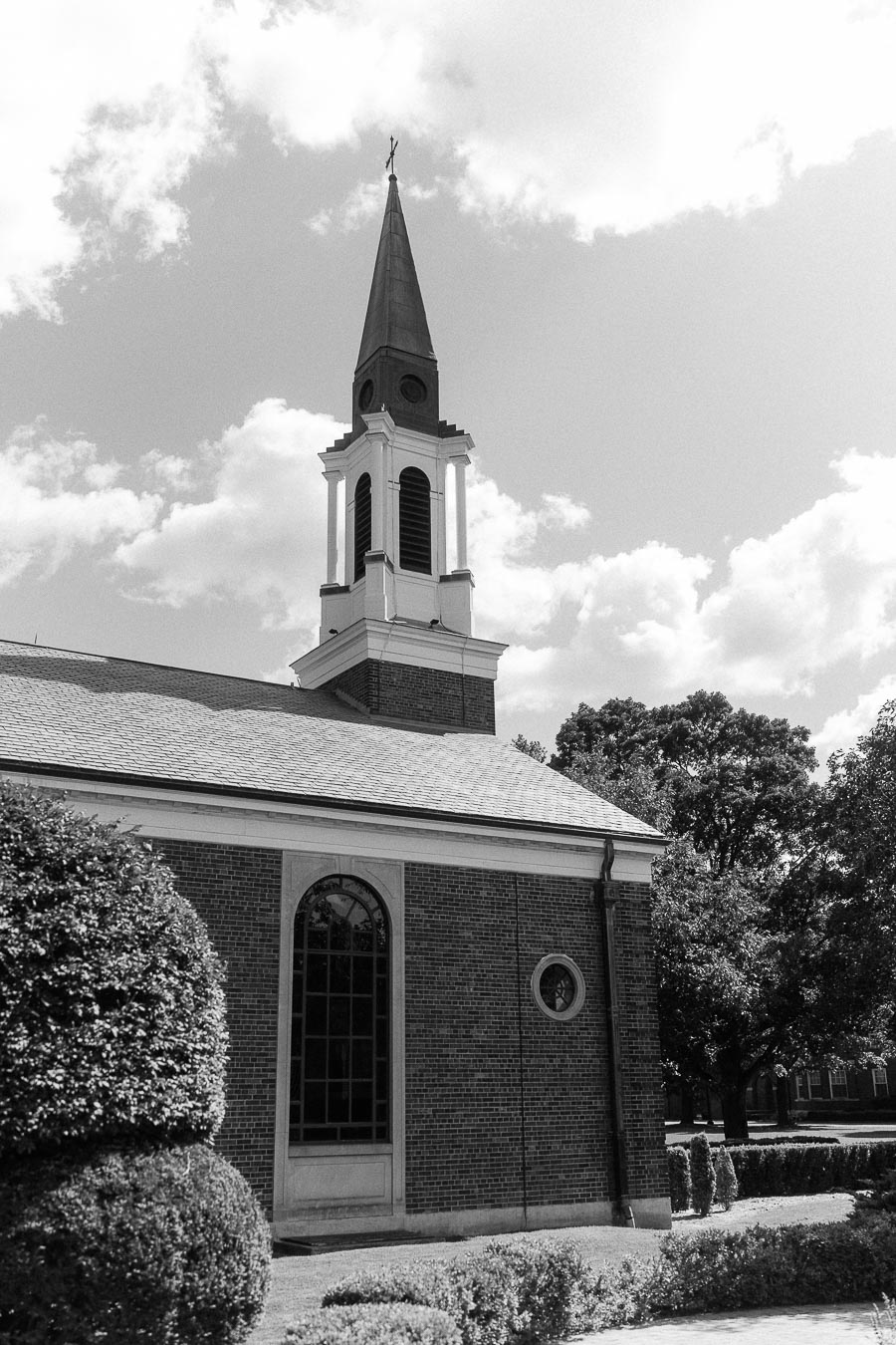 Black and white view of a brick chapel with a tall steeple, arched window, shrubs, and cloudy sky.