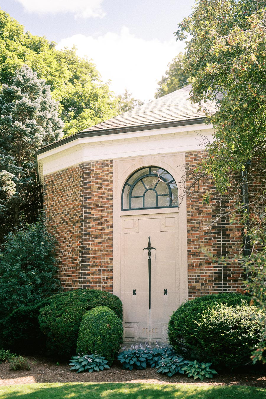 Brick garden building with an arched cream door framed by shrubs and summer trees in Aurora, Ontario.