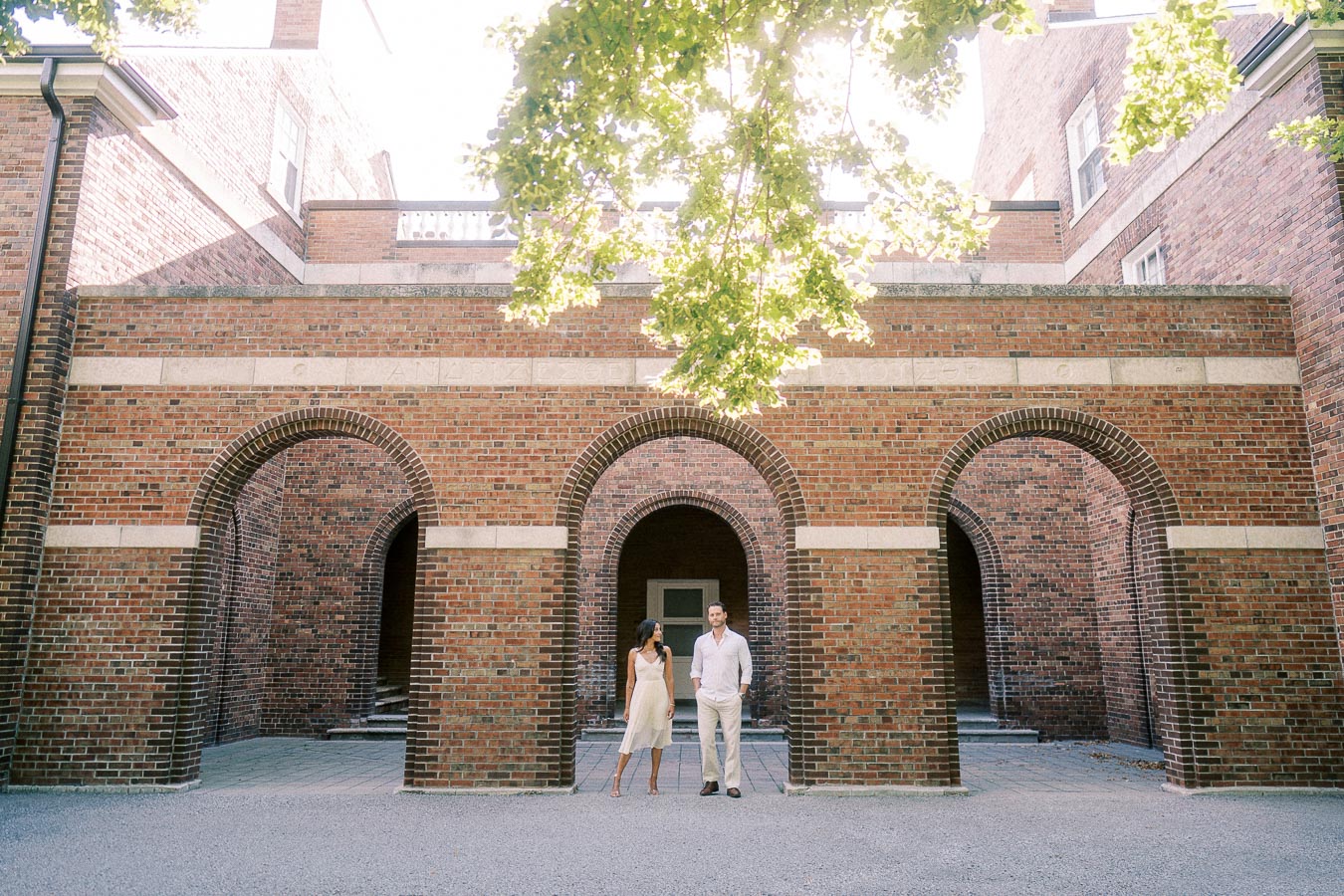 Rachel and Matt walk together beneath tall brick arches in a sunlit campus courtyard.