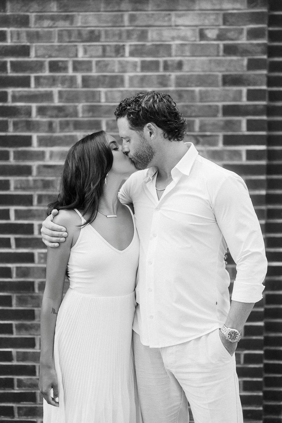 Rachel and Matt kiss while standing close together in front of a brick wall in Aurora, Ontario.