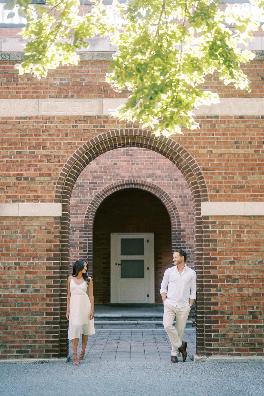 Rachel and Matt stand apart beneath a tall brick archway, smiling at each other in the summer light.