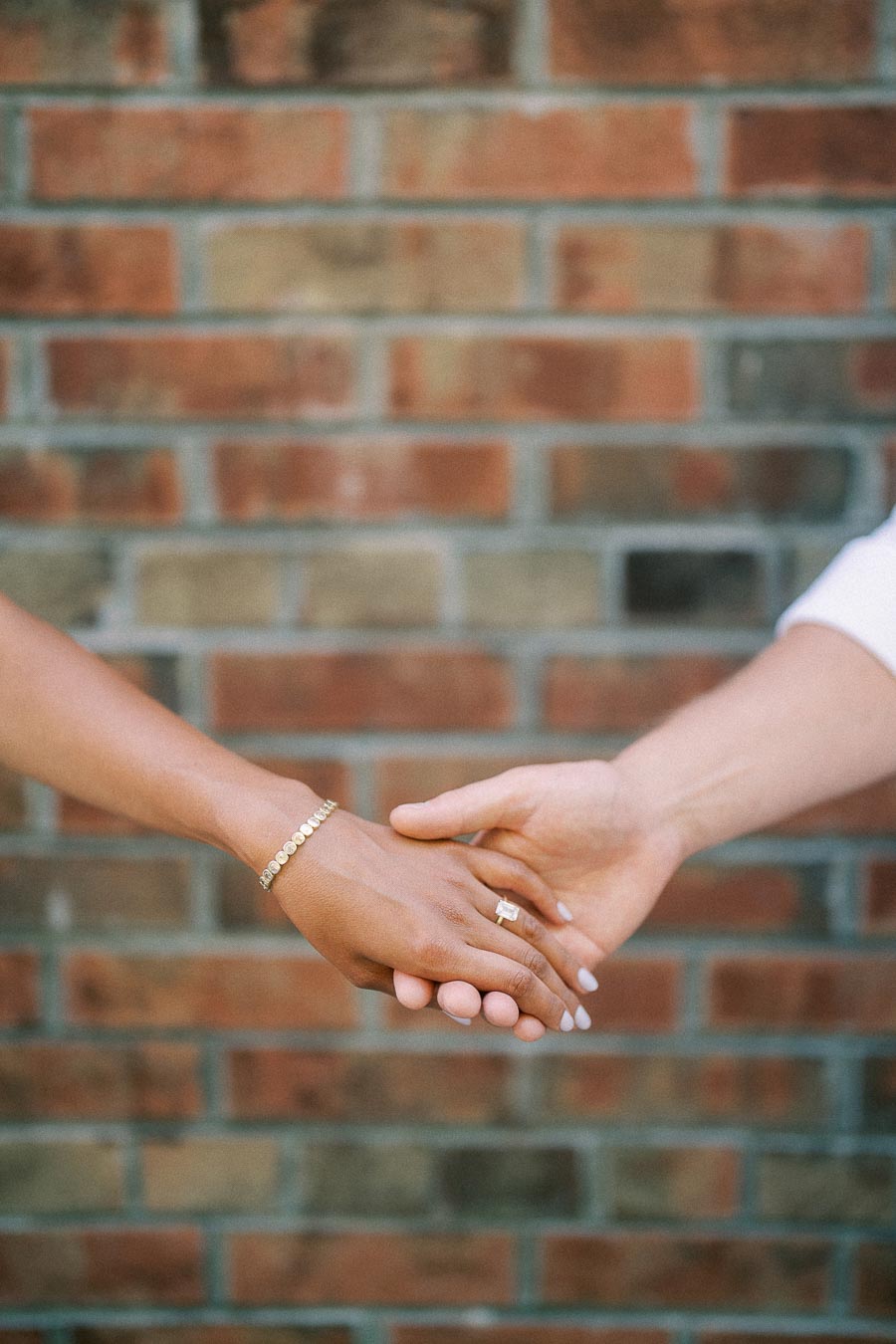 Rachel and Matt hold hands in front of a brick wall, with her engagement ring visible.