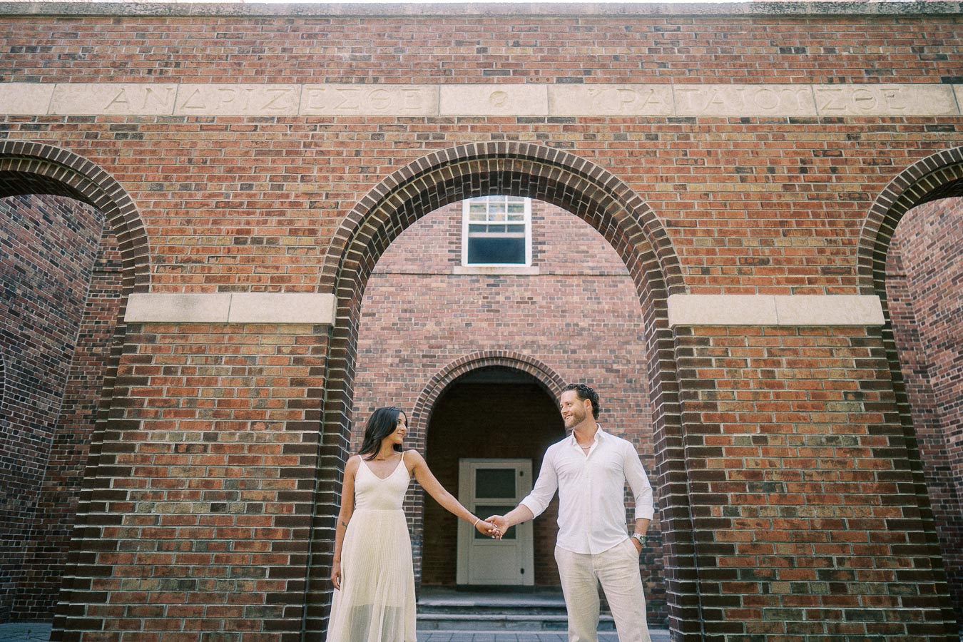 Rachel and Matt hold hands beneath tall brick arches at St. Andrew’s College in Aurora.