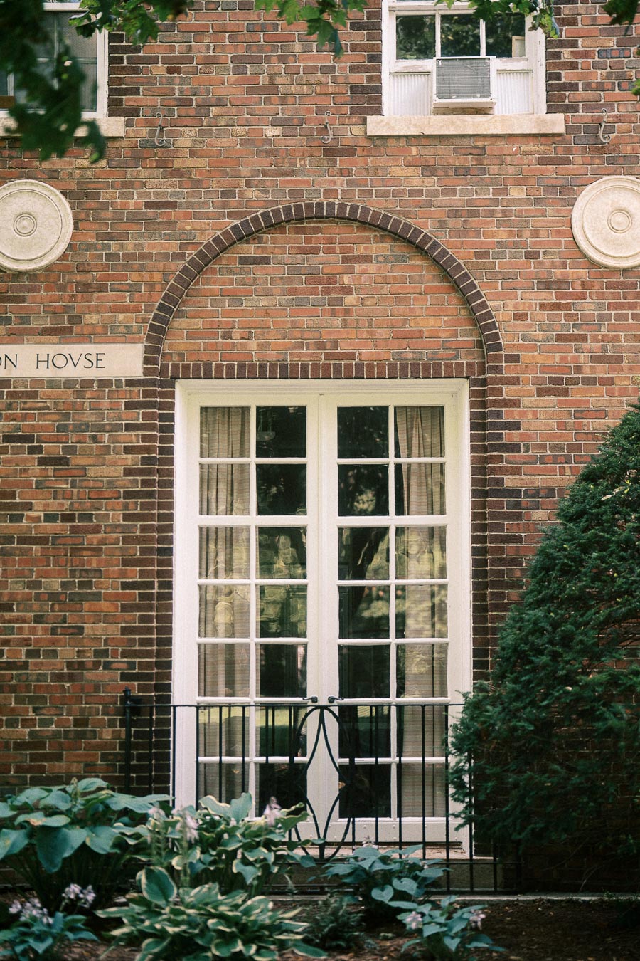 Brick building façade with white French doors, arched brickwork, garden plants, and leafy branches.