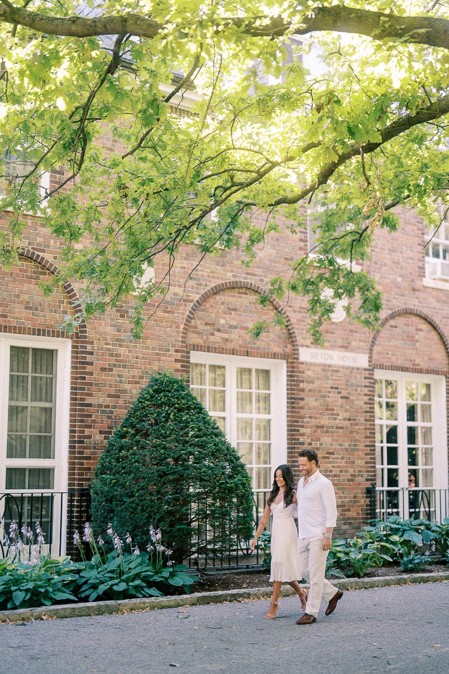 Rachel and Matt walk together past a brick school building beneath leafy green branches in Aurora, Ontario.